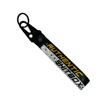 Front side of a black, white and yellow keychain with gritty design reading "Authentic Sh*tbox" – Motorev automotive keychain accessory
