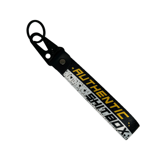 Front side of a black, white and yellow keychain with gritty design reading "Authentic Sh*tbox" – Motorev automotive keychain accessory